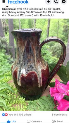 a brown and black vase sitting on top of a table next to pink flowers in the forest