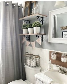 a white toilet sitting under a bathroom window next to a sink and shower curtain with potted plants on it