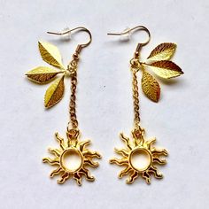 two gold earrings with leaves hanging from them on a white surface, one has a sun and the other is a leaf