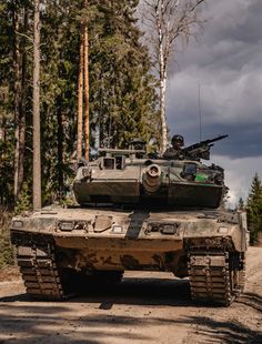 an army tank driving down a dirt road in the woods with trees and clouds behind it