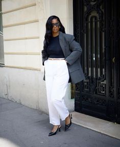 a woman standing in front of a building wearing white pants and a black top with a gray blazer