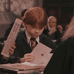 a young boy sitting at a table reading a book and holding a piece of paper