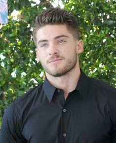 a man with a beard and black shirt posing for a photo in front of some bushes