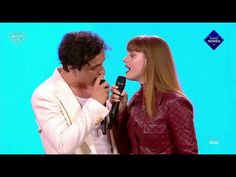 a man holding a microphone next to a woman singing into it's mouth while standing in front of a blue background