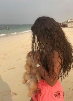 a woman holding a small dog on top of a sandy beach next to the ocean