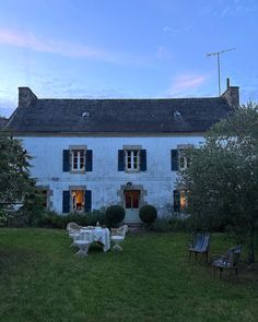 an old white house with blue shutters and tables in the front yard at dusk