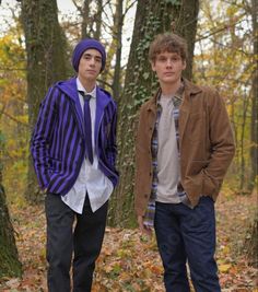 two young men standing next to each other in front of trees with leaves on the ground