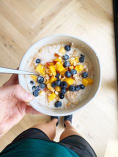 a person holding a bowl of oatmeal with blueberries and mangoes