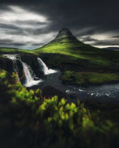 a waterfall in the middle of a lush green field next to a mountain under a cloudy sky