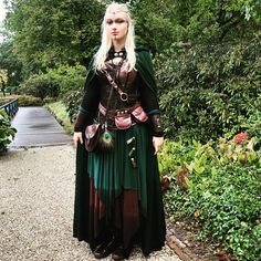 a woman dressed in green and black is standing on a path with trees behind her