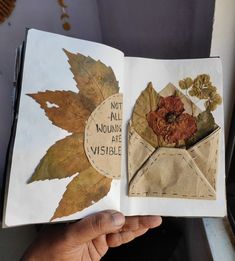 an open book with leaves and flowers on the pages is held in someone's hand