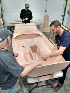 two men working on a wooden boat in a shop with another man looking at it