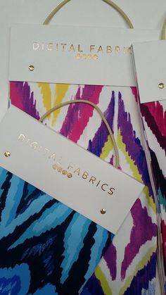 two tags are attached to the handles of some colorful fabric bags with gold lettering on them