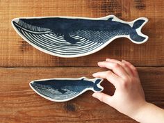 two blue whale shaped magnets on a wooden surface, one being held up by a person's hand