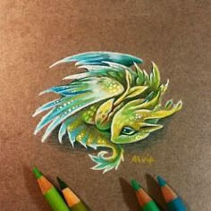 a drawing of a green dragon with blue wings on it's head and two colored pencils next to it