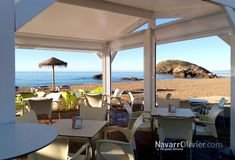 tables and chairs are set up under an umbrella on the beach with water in the background