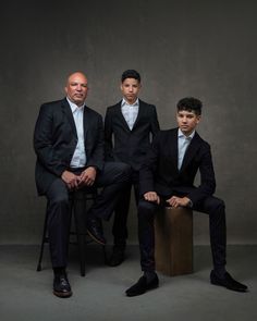 three men in suits are sitting on a stool