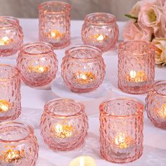 many pink glass candles are lit on a white table cloth with flowers in the background