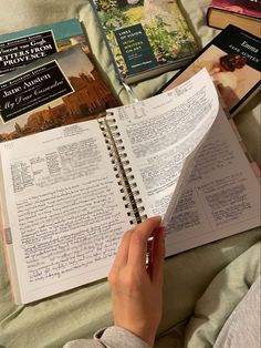 a person reading a book on top of a bed next to other books and papers