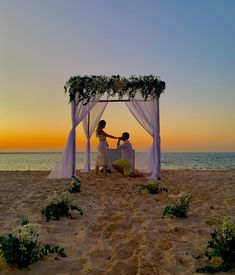 two people are sitting under a canopy on the beach at sunset, with an ocean view in the background