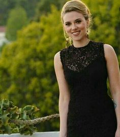 a woman in a black dress standing on a balcony next to a tree and bushes