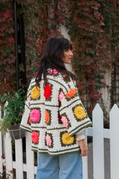 a woman standing in front of a white fence wearing a crocheted jacket with flowers on it