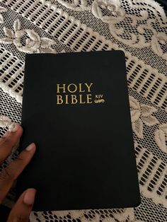 a hand holding a black bible on top of a white lace tablecloth with gold lettering