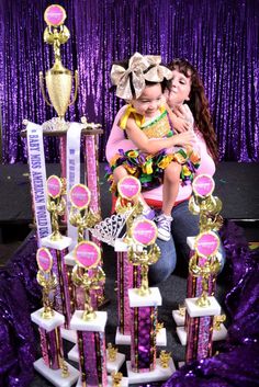 a woman holding a small child in front of trophies on a purple tablecloth covered stage