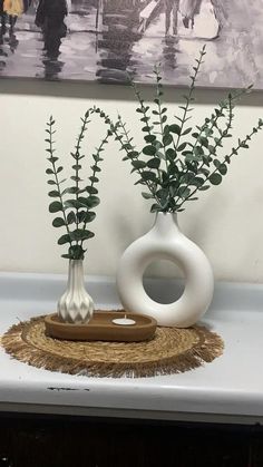 two white vases with plants in them sitting on a table next to a painting