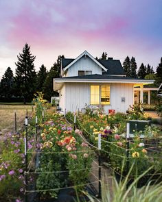 a white house surrounded by lots of flowers and plants in front of it at dusk