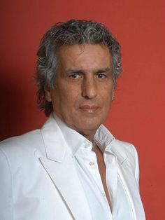 a man with grey hair wearing a white suit and posing for a photo in front of a red wall