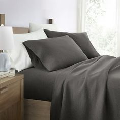 a bed with grey sheets and pillows in a bedroom next to a window, lamp and nightstand