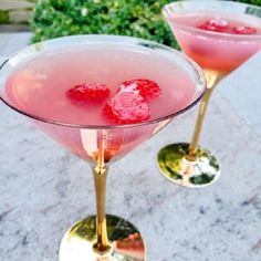 two martinis with raspberries sit on a marble table in front of bushes