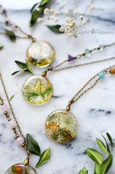 four necklaces with flowers and leaves on them sitting on a marble counter top, surrounded by greenery