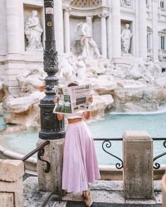 a woman in a pink dress reading a newspaper near a fountain with statues on it