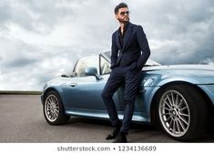 a man sitting on the hood of a blue sports car with clouds in the background