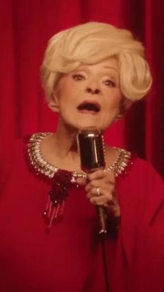 a woman with blonde hair holding a microphone in front of red curtained walls and looking surprised