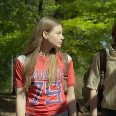 two young women walking in the woods with backpacks on their back and one wearing a red shirt