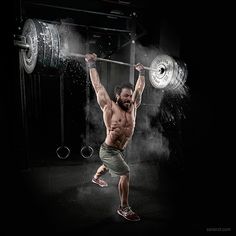 a shirtless man lifts a barbell over his head while standing in the dark