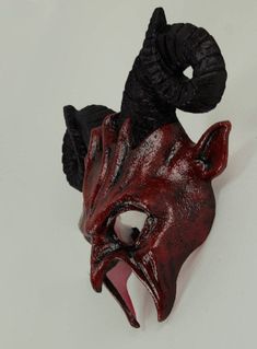 a red and black mask with its mouth open on a white surface, next to an object that looks like a demon's head