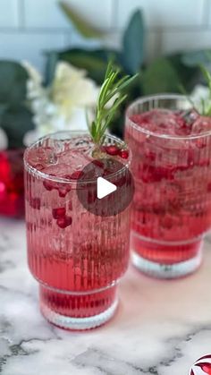 two glasses filled with red liquid and topped with a sprig of rosemary sitting on top of a marble counter