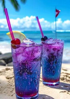 two glasses filled with purple liquid sitting on top of a sandy beach next to the ocean