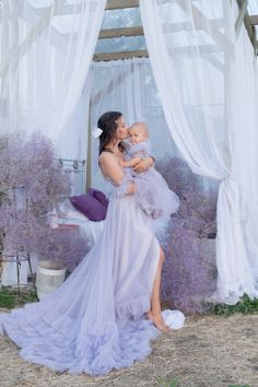 a woman holding a baby in her arms while standing next to a window with sheer curtains