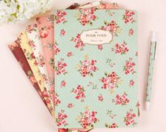 three notebooks with flowers on them next to a pen and flower arrangement in the background