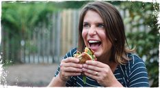 a woman eating a sandwich with her mouth wide open and smiling at the camera while sitting outside