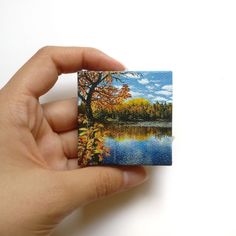 a hand holding up a small piece of paper with a lake and trees on it