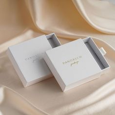 two white boxes sitting on top of a satin covered tablecloth with gold lettering that reads, dancaew jewelry