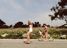 two women in bathing suits on skateboards and one riding a bike with her arms up