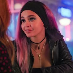 a woman with pink hair wearing a black leather jacket and headband talking to another woman