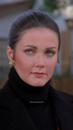 a close up of a person wearing a black coat and gold earrings with a house in the background